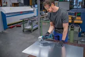 Turvavarusteita käyttävä henkilö leikkaa metallilevyä akkukäyttöisellä sähkötyökalulla.