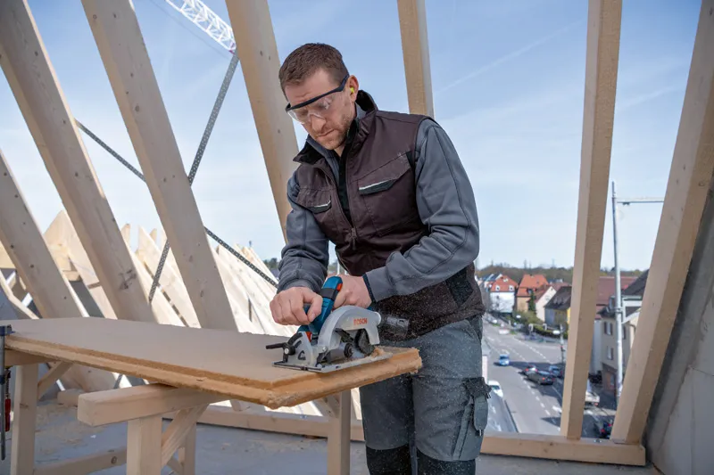 Turvavarusteita käyttävä henkilö sahaa puulautaa pyörösahalla rakennustyömaalla.