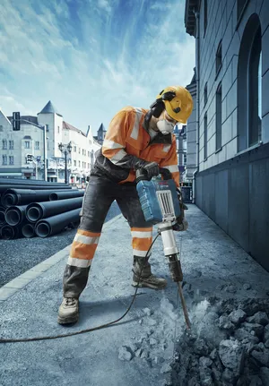 Turvavarusteita käyttävä henkilö rikkoo betonipäällystettä purkuvasaralla.