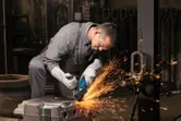 Turvavarusteita käyttävä henkilö hioo metallipintaa kulmahiomakoneella.