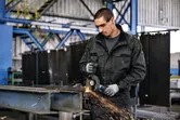 Turvavarusteita käyttävä henkilö hioo metallia kulmahiomakoneella työpajassa.