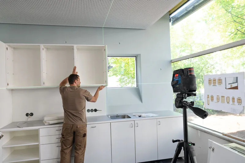 Henkilö kohdistaa seinäkaappeja laserilla vaaitustyökalulla keittiöasennuksessa.