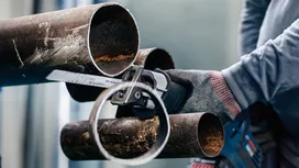 Turvavarusteita käyttävä henkilö leikkaa suurta ruostunutta metalliputkea edestakaisin liikkuvalla sahalla.