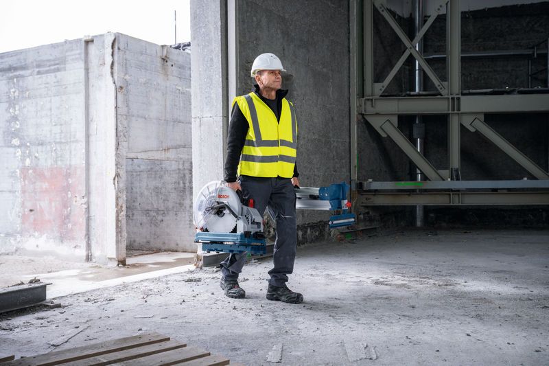 Turvavarusteita käyttävä henkilö kantaa metallin katkaisusahaa keskeneräisessä rakennuksessa.