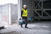 Turvavarusteita käyttävä henkilö kantaa metallin katkaisusahaa keskeneräisessä rakennuksessa.