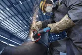 Turvavarusteita käyttävä henkilö hioo metalliputkea kulmahiomakoneella.