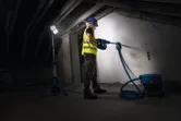Turvavarusteita käyttävä henkilö poraa seinään johdottoman työmaavalon alla.