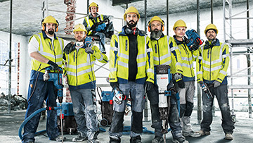 Seitsemän työntekijää, keltaiset kypärät, Bosch-työkalut, rakennustyömaa.