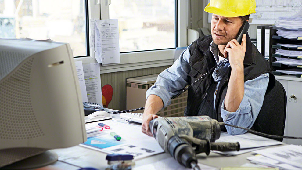 Työmies puhuu puhelimeen työpöydällä, jossa on porakone ja asiakirjoja.