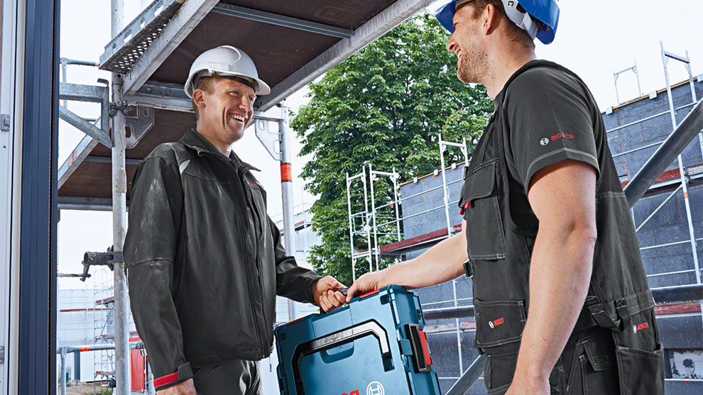 Kaksi miestä hymyilee ja vaihtaa Bosch-työkalulaukkua rakennustyömaalla.