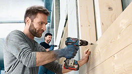 Mies käyttää Bosch-akkuporakoneita puupinnalla, taustalla toinen henkilö.
