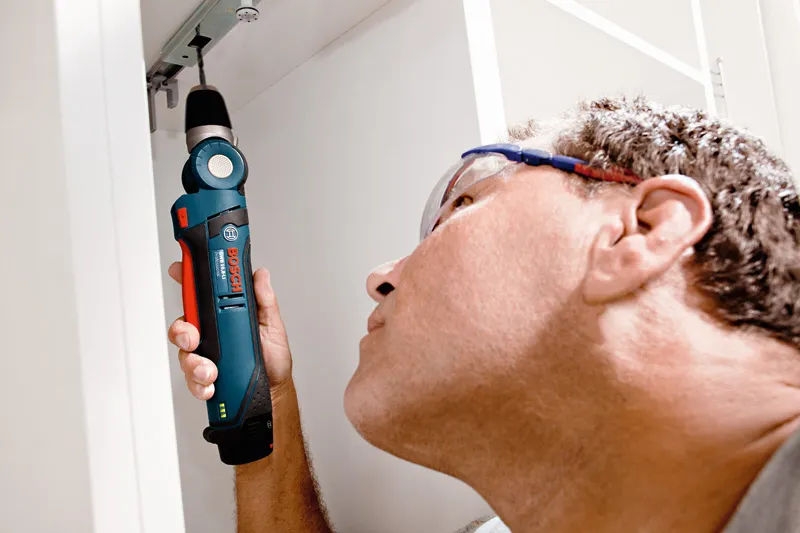 Une personne portant un équipement de sécurité utilise une perceuse d’angle sans fil pour travailler à l’intérieur d’une armoire.