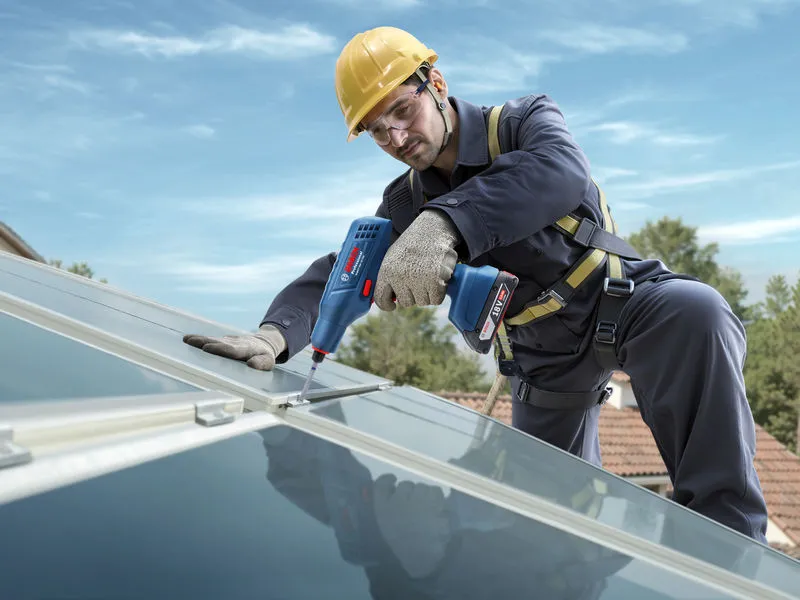 Une personne portant un équipement de sécurité serre un boulon sur un panneau solaire sur le toit avec un outil électrique.