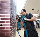 Une personne portant un équipement de sécurité utilise une perceuse avec un système d'extraction de poussière sur un mur de briques.