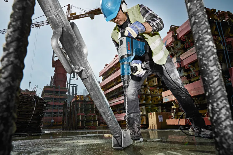 Un ouvrier portant un équipement de sécurité utilise un marteau perforateur pour percer une base en béton sur un chantier de construction.