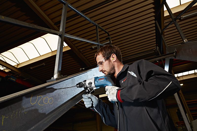 Une personne portant un équipement de sécurité perce une poutre métallique sur un site industriel.