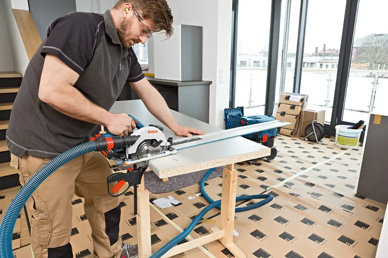 Une personne portant un équipement de sécurité coupe un panneau de bois avec une scie circulaire sans fil sur un établi.