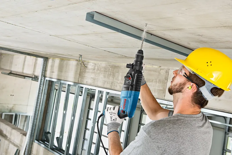 Une personne portant un équipement de sécurité perce un plafond en béton avec un marteau perforateur.