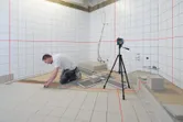 Une personne installe des carreaux de sol dans une salle de bain, guidée par un outil de nivellement laser sur un trépied.