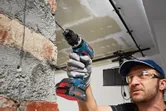 Une personne portant un équipement de sécurité perce un mur de briques avec une perceuse électrique sans fil.