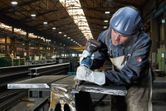 Une personne portant un équipement de sécurité perce une poutre métallique dans une usine.