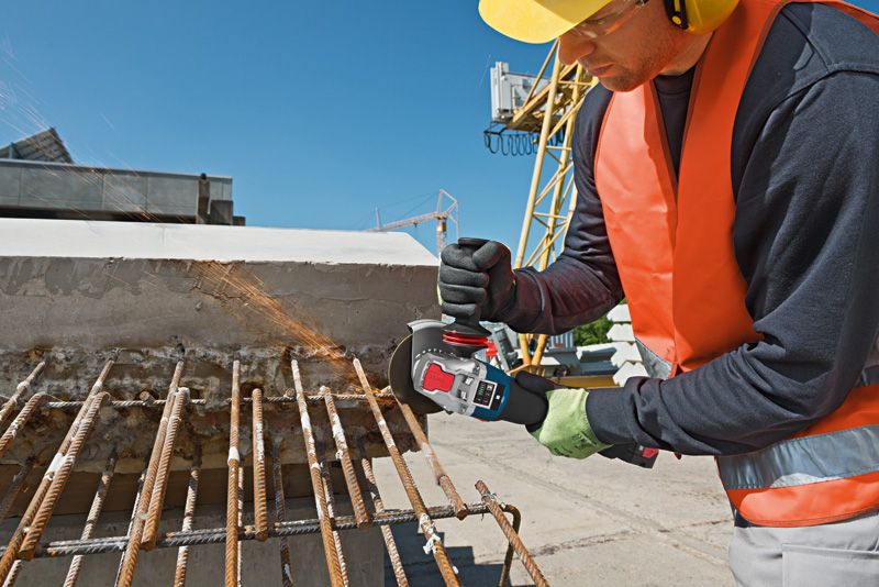 Une personne portant un équipement de sécurité coupe des barres d'armature rouillées avec une meuleuse d'angle sur un chantier de construction.