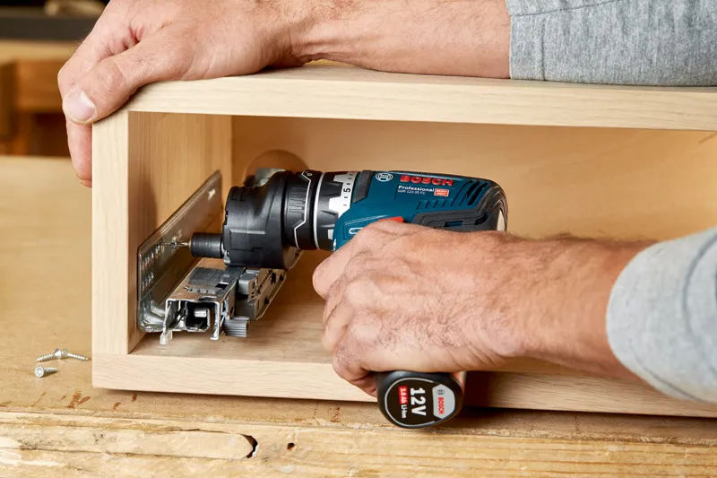 Personne utilisant une perceuse sans fil pour installer une glissière de tiroir en métal à l'intérieur d'une boîte en bois.