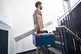 Une personne porte une mallette à outils et un mesureur d’angle en montant les escaliers.