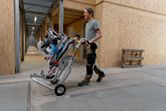 Une personne portant un équipement de sécurité transporte une scie à onglets sans fil sur un support à roulettes.