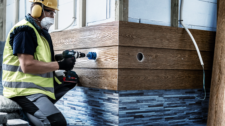 Une personne portant un équipement de sécurité utilise une perceuse électrique munie d'une scie cloche sur un mur en bois.