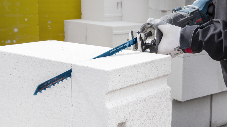 Une personne portant un équipement de sécurité utilise une scie alternative pour couper un bloc de béton blanc.