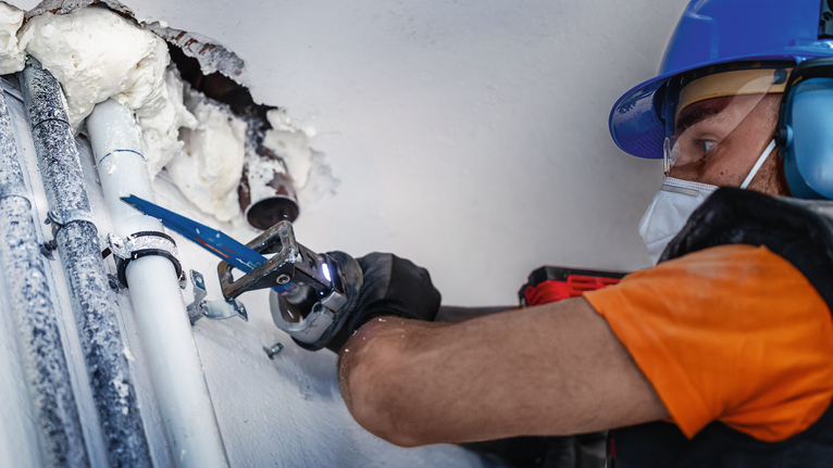 Une personne portant un équipement de sécurité utilise une scie alternative pour couper un tuyau sur un mur.
