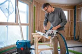 Une personne portant un équipement de sécurité coupe du bois avec une scie circulaire dans une pièce en construction.