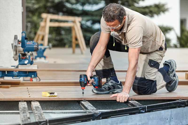 Une personne portant un équipement de sécurité utilise une perceuse électrique pour fixer des planches de terrasse à l'extérieur.