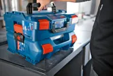Personne soulevant un système de mallette de transport bleue empilée avec des loquets orange sur une table.