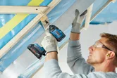 Une personne portant un équipement de sécurité utilise une perceuse sans fil pour fixer une poutre métallique au bois.
