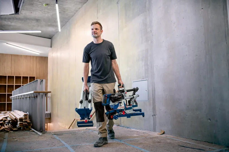 Une personne transporte une scie à onglets sans fil et son trépied sur un chantier de construction moderne.