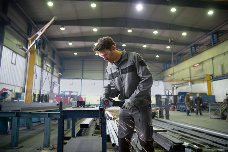 Une personne portant un équipement de sécurité broie du métal, des étincelles jaillissent dans un grand atelier industriel.