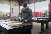 Un ouvrier portant un équipement de sécurité broie du métal sur un grand établi dans une usine.