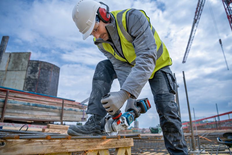 Une personne portant un équipement de sécurité coupe des barres d'armature avec une meuleuse d'angle sans fil sur un chantier de construction.