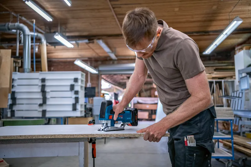 Une personne portant un équipement de sécurité coupe une planche de bois avec une scie sauteuse dans un atelier.