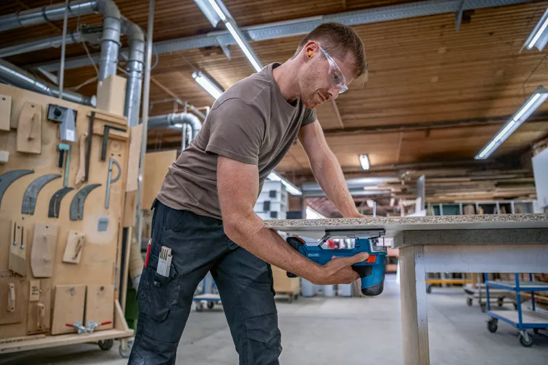 Une personne portant un équipement de sécurité coupe une planche de bois avec une scie électrique dans un atelier.