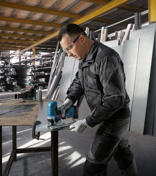 Une personne portant un équipement de sécurité coupe de la tôle avec une scie sauteuse sans fil dans un atelier.