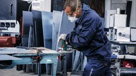 Une personne portant un équipement de sécurité découpe une tôle de métal à l'aide d'une scie électrique dans un atelier.