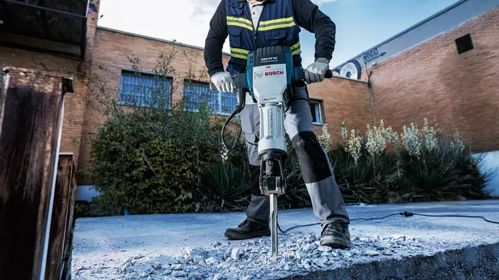 Une personne portant un équipement de sécurité brise du béton avec un marteau de démolition à l'extérieur.