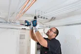 Une personne portant un équipement de sécurité perce un plafond pour installer un conduit électrique.