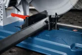 Scie à métaux coupant un tuyau métallique carré sur une surface de travail bleue.