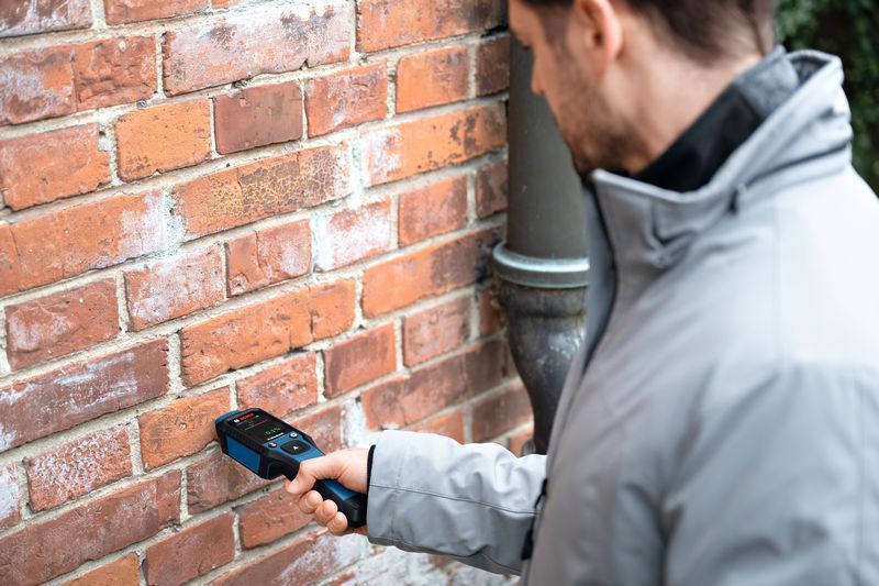 Une personne scanne un mur de briques avec un humidimètre.