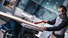 Une personne portant un équipement de sécurité guide une pièce de bois à travers une scie circulaire dans un atelier.
