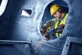 Une personne portant un équipement de sécurité meule du métal avec une meuleuse d'angle à l'intérieur d'une grande structure.